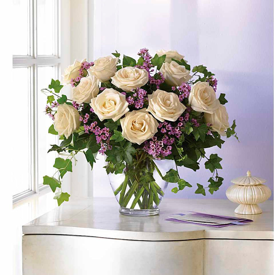 Bouquet of white roses with purple flowers in a clear vase on a table.
